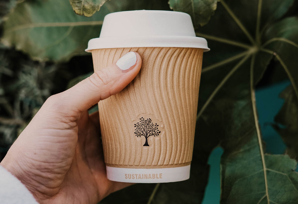 hand holding disposable coffee cup with the word sustainable in front of leaves