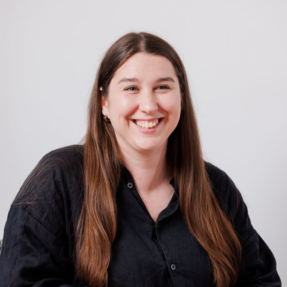 Headshot of Rachel, a white woman in her 30s with long brown hair. She's wearing a black blouse and smiling off camera.